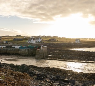 Portnaguran: A coastal fishing village with white hebridean houses in the distance, and the sea, shoreline and a pile of creels in the foreground.