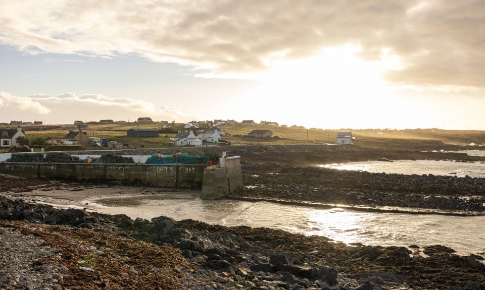 Portnaguran: A coastal fishing village with white hebridean houses in the distance, and the sea, shoreline and a pile of creels in the foreground.