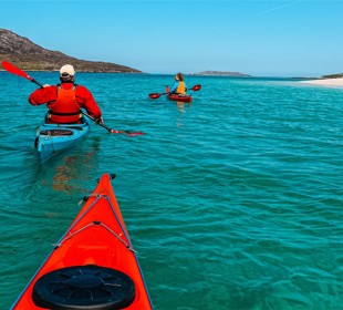 Two people in red kayaks paddle on turquoise water near a sandy beach with rocky hills and a clear blue sky.