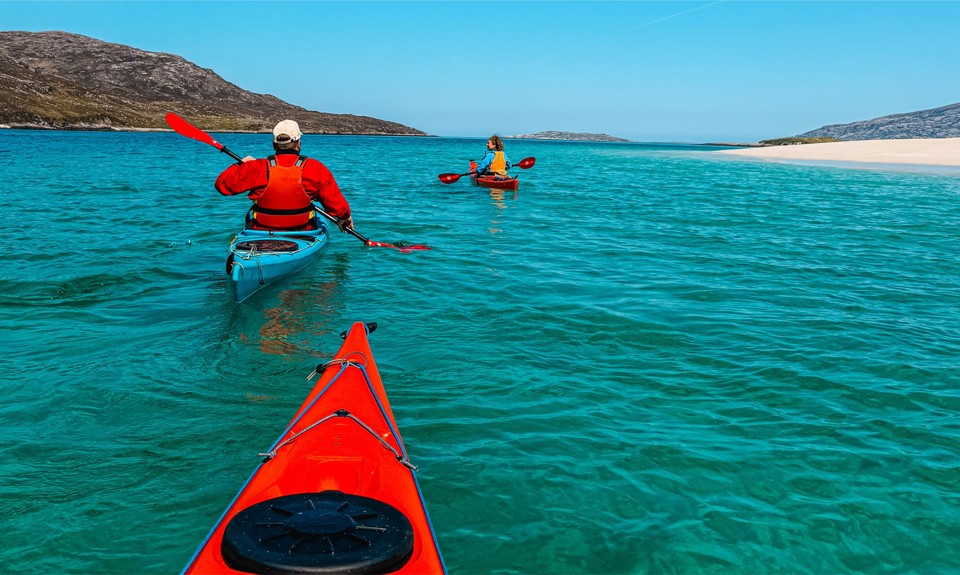 Two people in red kayaks paddle on turquoise water near a sandy beach with rocky hills and a clear blue sky.