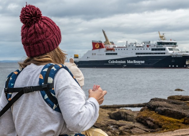 A person holding a child looks toward a Caledonian MacBrayne ferry docked near the shore on a cloudy day.