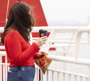 A female with long brown hair, wearing a red sweater, is standing on the deck holding a black cup with the CalMac logo. 