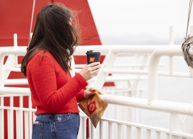 A female with long brown hair, wearing a red sweater, is standing on the deck holding a black cup with the CalMac logo. 
