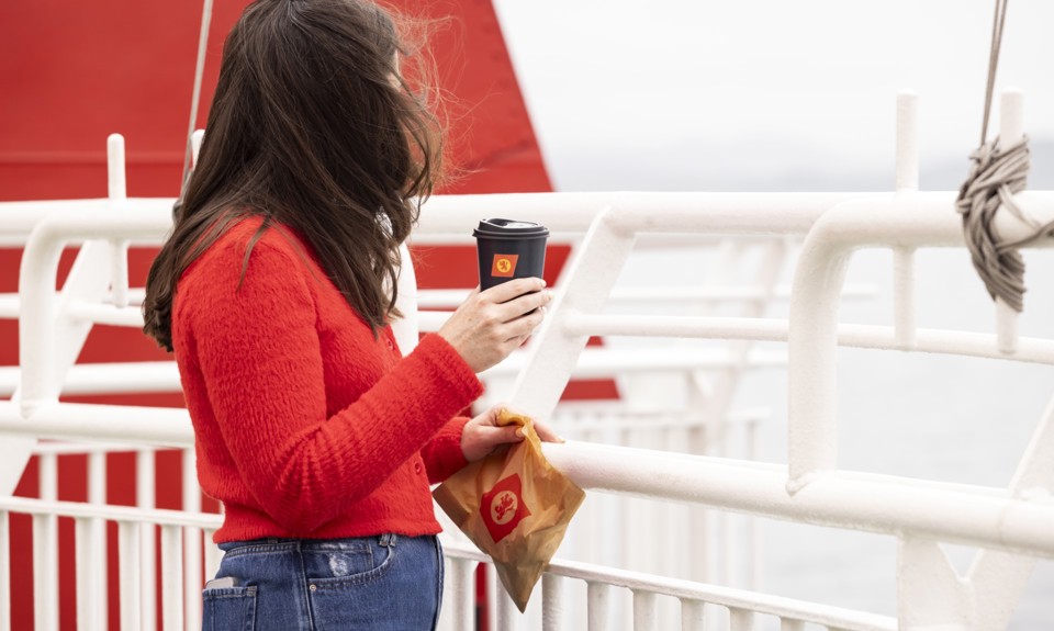 A female with long brown hair, wearing a red sweater, is standing on the deck holding a black cup with the CalMac logo. 