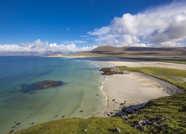 A view of the long golden sandy beach of Luskentyre, with blue sky and clear water rolling in.