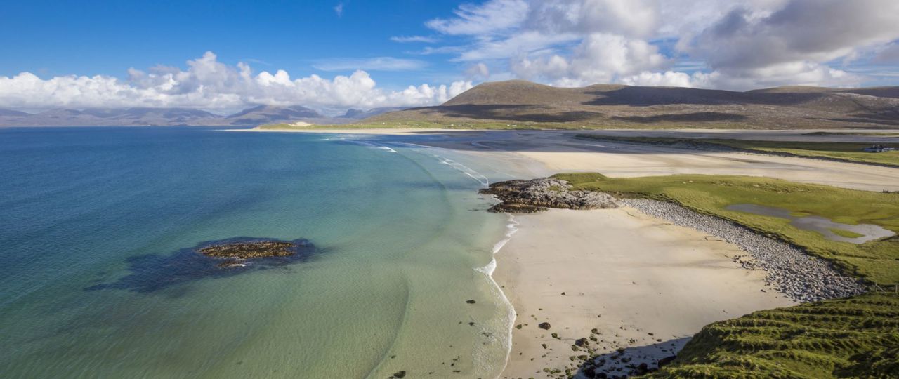 A view of the long golden sandy beach of Luskentyre, with blue sky and clear water rolling in.