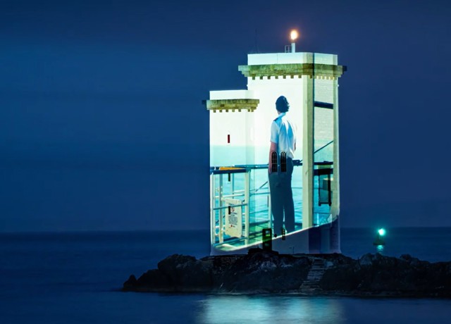 A projected image of a CalMac Officer, onto a lighthouse, while everything around it is in darkness.