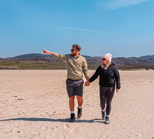 Two people walk hand-in-hand on sandy beach; one points toward horizon. Hills and house visible in background.