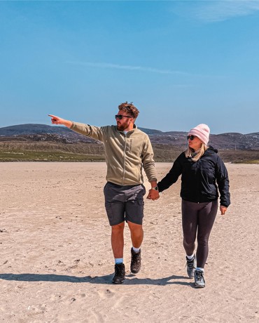 Two people walk hand-in-hand on sandy beach; one points toward horizon. Hills and house visible in background.