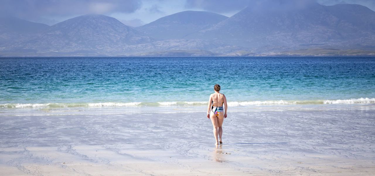 A woman walking to the blue waters edge across a sandy beach with hills in the background