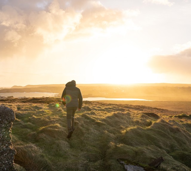 Person walking on grassy coastal hill at sunset overlooking sea and islands.