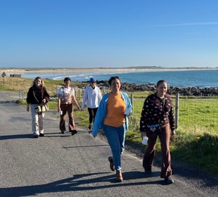 Five people walking along the paved coastal path with the sea and rocky shoreline to the right, under a clear blue sky, surrounded by green grass.