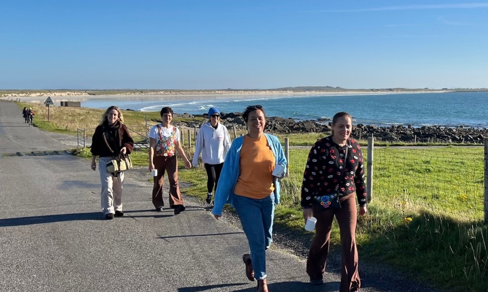 Five people walking along the paved coastal path with the sea and rocky shoreline to the right, under a clear blue sky, surrounded by green grass.