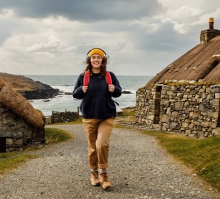 Female with yellow hat and red backpack walking through an old stone village with the sea in the background