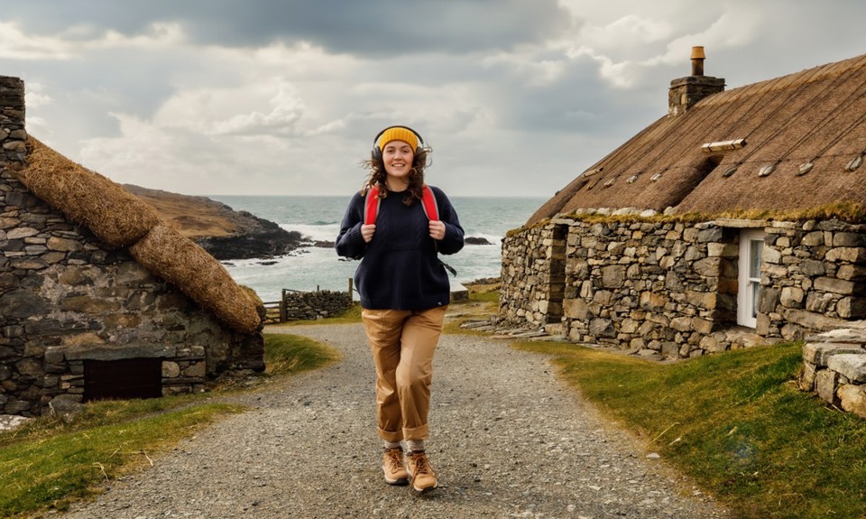 Female with yellow hat and red backpack walking through an old stone village with the sea in the background
