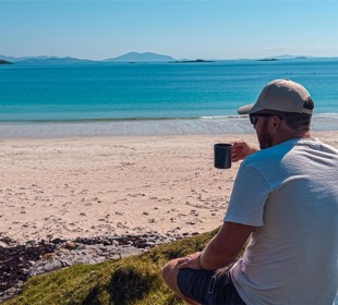 Man in white shirt and cap sits on grassy hill with a mug, overlooking a turquoise beach and distant mountains.