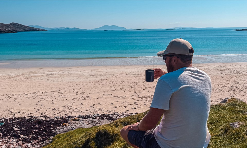 Man in white shirt and cap sits on grassy hill with a mug, overlooking a turquoise beach and distant mountains.