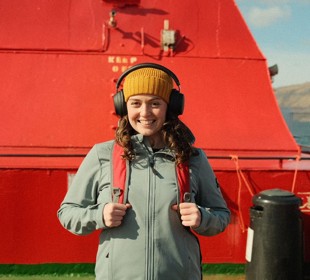 Brunette female smiling at camera, wearing a yellow hat and black over ear headphones, on an upper deck of a ferry beside a red funnel