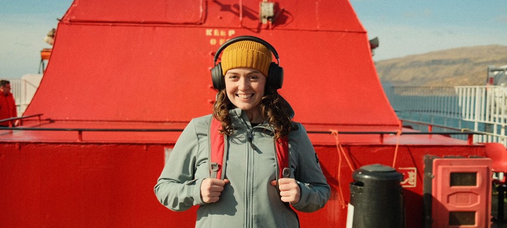 Brunette female smiling at camera, wearing a yellow hat and black over ear headphones, on an upper deck of a ferry beside a red funnel