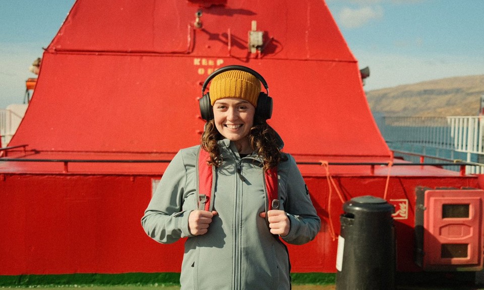 Brunette female smiling at camera, wearing a yellow hat and black over ear headphones, on an upper deck of a ferry beside a red funnel