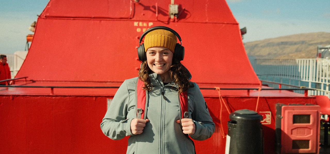 Brunette female smiling at camera, wearing a yellow hat and black over ear headphones, on an upper deck of a ferry beside a red funnel