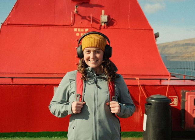 Brunette female smiling at camera, wearing a yellow hat and black over ear headphones, on an upper deck of a ferry beside a red funnel