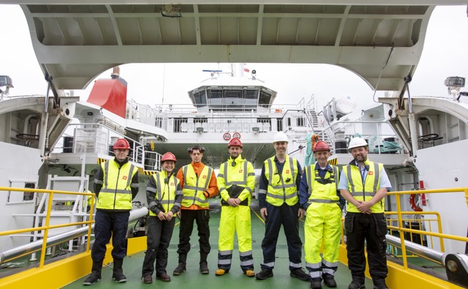 Crew members in high‑visibility gear standing on a ferry deck with the vessel’s bridge visible behind them.