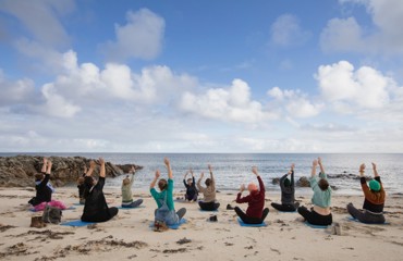 A group of people sitting on yoga mats on a sandy beach, facing the ocean. They are performing a yoga pose with their arms raised towards the sky. 