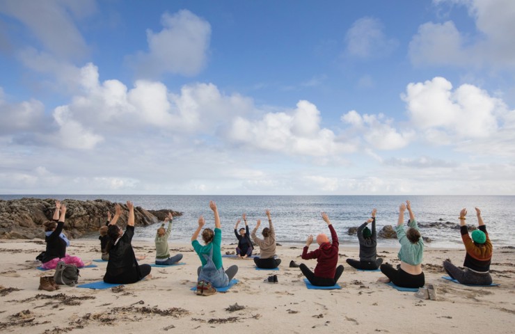 A group of people sitting on yoga mats on a sandy beach, facing the ocean. They are performing a yoga pose with their arms raised towards the sky. 