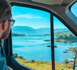 Man with glasses looks out a vehicle window at a scenic view of a lake and mountains.