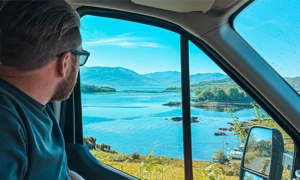 Man with glasses looks out a vehicle window at a scenic view of a lake and mountains.