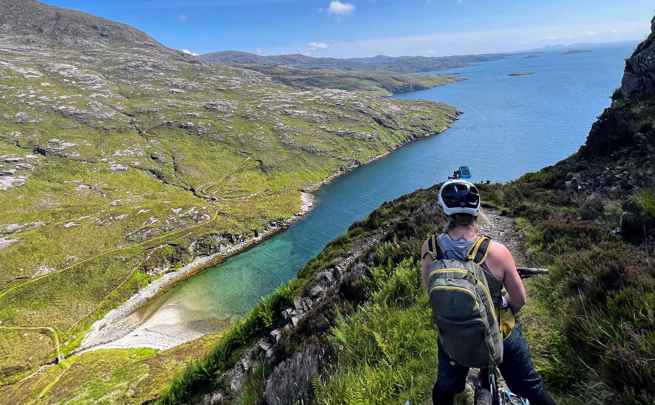 Cyclist on a high trail looking down at the green and rocky coastline.