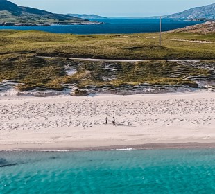 Aerial view of a beach with turquoise water, white sand, grassy dunes, and two people walking along the shore.