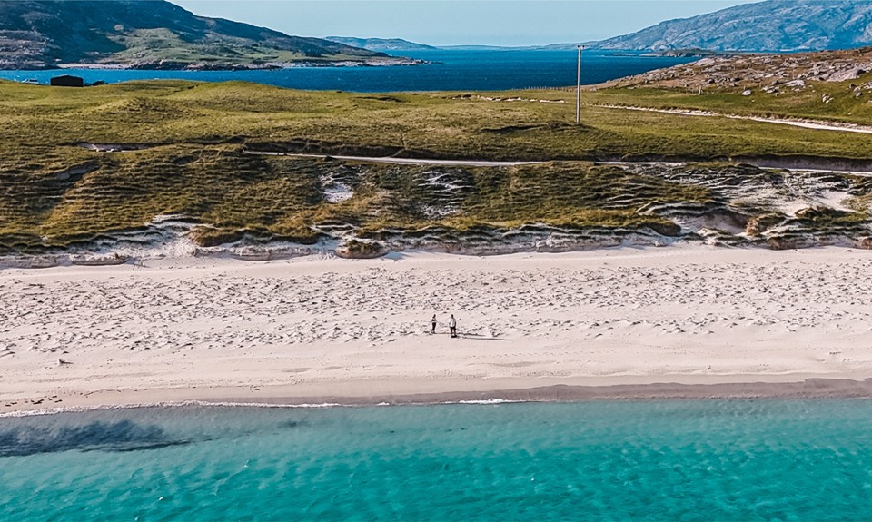 Aerial view of a beach with turquoise water, white sand, grassy dunes, and two people walking along the shore.
