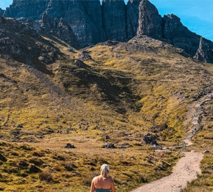 Person with blonde hair walks on a dirt path toward a rocky mountain under a clear blue sky, surrounded by grassy terrain.