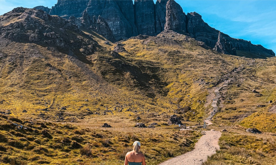Person with blonde hair walks on a dirt path toward a rocky mountain under a clear blue sky, surrounded by grassy terrain.