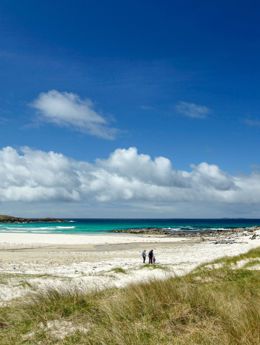 A family walking across a golden sandy beach on North Uist with dunes in the forground and blue sea and sky in the background.