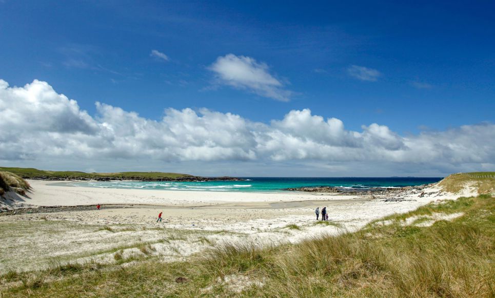 A family walking across a golden sandy beach on North Uist with dunes in the forground and blue sea and sky in the background.