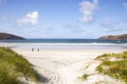 A couple walking across a sandy beach, under a blue sky to the water on a sunny day