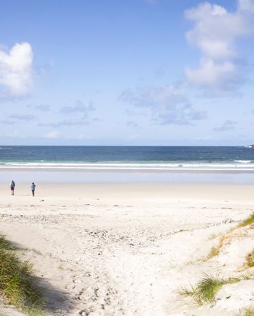 A couple walking across a sandy beach, under a blue sky to the water on a sunny day