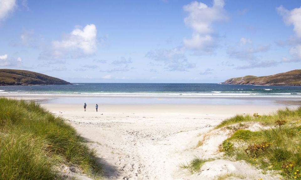 A couple walking across a sandy beach, under a blue sky to the water on a sunny day