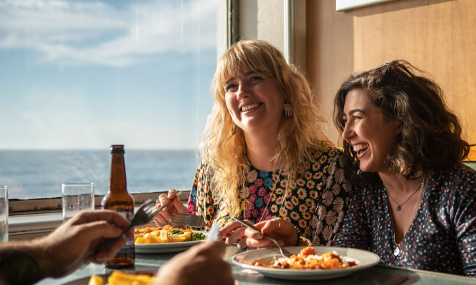 Two women laughing and eating food at a window seat looking out to sea