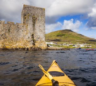 A yellow canoe sailing towards Kisimul Castle in a bay, with Castlebay village along the coast. Barra.