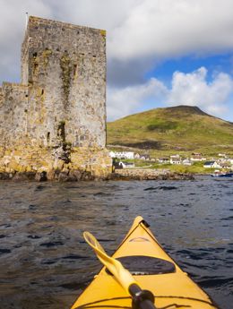 A yellow canoe sailing towards Kisimul Castle in a bay, with Castlebay village along the coast. Barra.