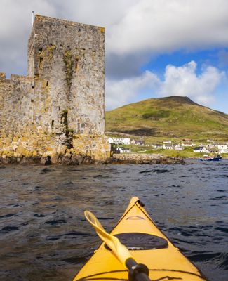 A yellow canoe sailing towards Kisimul Castle in a bay, with Castlebay village along the coast. Barra.