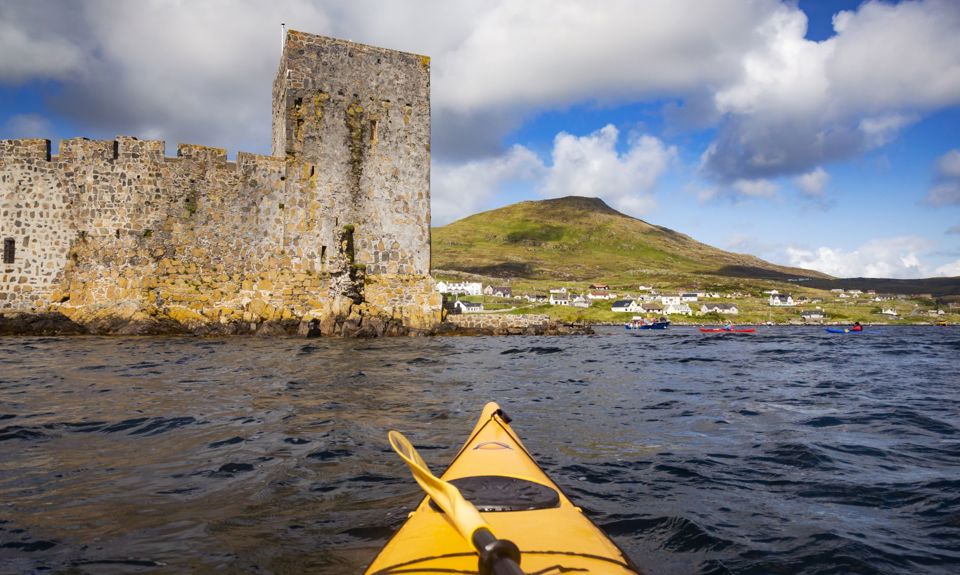 A yellow canoe sailing towards Kisimul Castle in a bay, with Castlebay village along the coast. Barra.