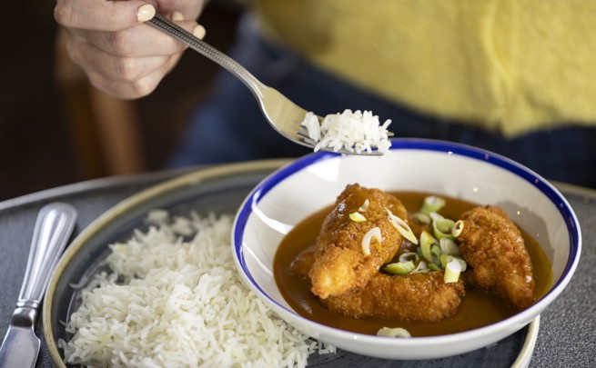 A person is holding a fork with white rice over a bowl of katsu curry garnished with chopped green onions. The bowl is placed on a plate.