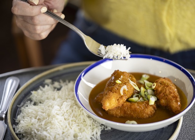 A person is holding a fork with white rice over a bowl of katsu curry garnished with chopped green onions. The bowl is placed on a plate.
