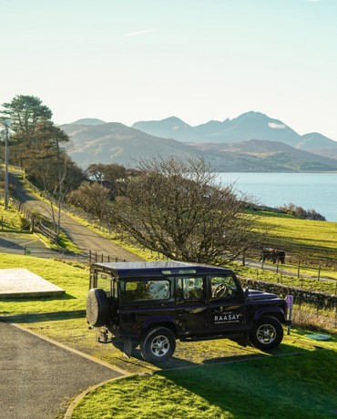 A dark 4x4 vehicle parked on a grassy area overlooking a loch, rolling hills, and sunlit mountains.