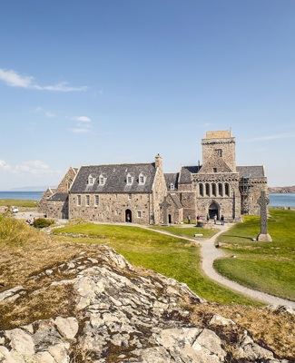 The ancient stone building of Iona Abbey taking from a rocky hilltop with the costline and water in the background.